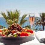A white plate with a Nicoise salad on it, topped with plenty of shredded fish, alongside a champagne glass filled with a pink Kir Royale cocktail. Behind, you can see the sea and two palm trees.