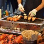 A person grilling lobster tails in the market, on a small rectangular barbecue