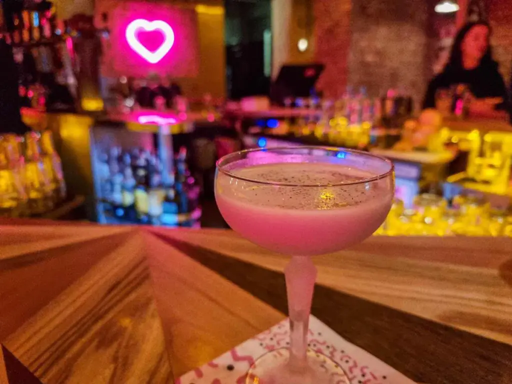 A Martini glass with a pink drink in it. Behind you can see blurred a pink neon heart on the wall