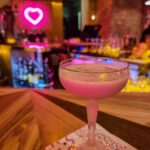A Martini glass with a pink drink in it. Behind you can see blurred a pink neon heart on the wall