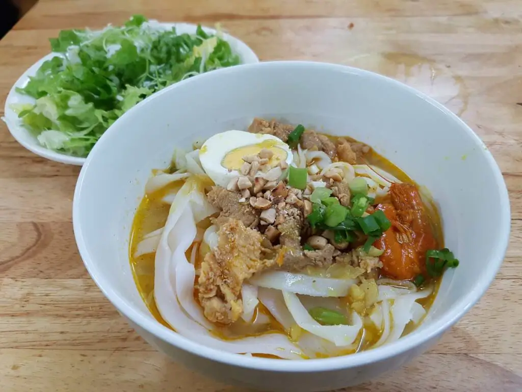 A white bowl with a soup topped with flat white noodles, fried pork, half an egg, red prawns, peanuts and spring onions. Behind there is a bowl with fresh salad.