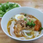A white bowl with a soup topped with flat white noodles, fried pork, half an egg, red prawns, peanuts and spring onions. Behind there is a bowl with fresh salad.