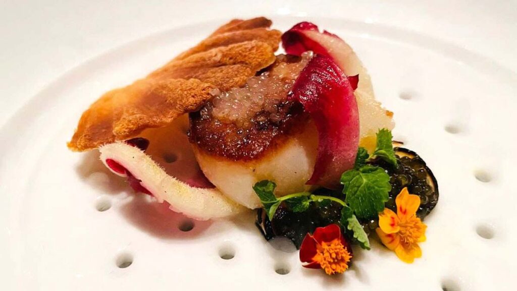 a scallop on a bed of red caviar, topped with white caviar, and a golden biscuit. The plate is decorated with edible orange marigolds.
