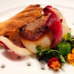 a scallop on a bed of red caviar, topped with white caviar, and a golden biscuit. The plate is decorated with edible orange marigolds.