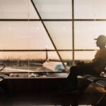 A man wearing a yellow tshirt, black jeans and a black cap, sitting down at a gate, inside the airport. The sun is setting so the light is very warm. At the gate, through the windows, you can see a white plane