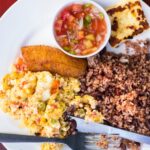 A white plate with beans and rice on it, scrambled eggs, fried plantain, fried cheese, and a tomato based sauce in a white ramekin.