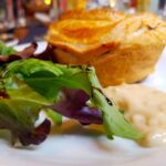 A gin infused pie with golden flaky pastry, on a white plate with a white sauce and leafy salad on the side.