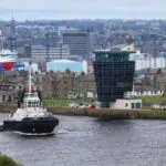 A coastguard ship sailing on the river in Aberdeen. Next to the river there are old houses. Behind them, there are a few sky scrapers and industrial buildings. Through them, you can see other old houses and churches in the city.
