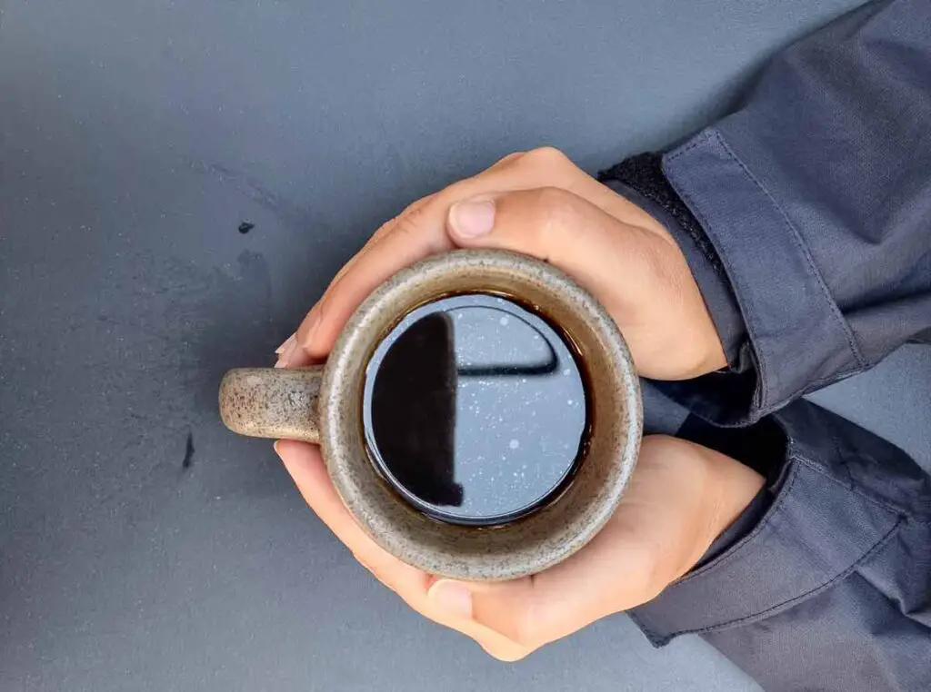 A close-up of my hands holding a cup of black coffee. The coffee is in a grey earth-ware mug. You can see the sleeves of my jacket, which is black.