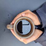A close-up of my hands holding a cup of black coffee. The coffee is in a grey earth-ware mug. You can see the sleeves of my jacket, which is black.