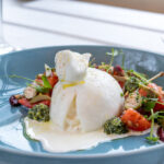 A buratta cheese on on a blue plate. On the right side there are chopped cherry tomatoes and rocket salad in a green pesto salsa.