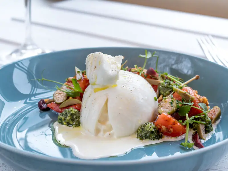 A buratta cheese on on a blue plate. On the right side there are chopped cherry tomatoes and rocket salad in a green pesto salsa.