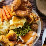 A large white plate with a Sunday roast on it. There are slices of beef, roast potatoes, a Yorkshire pudding, carrot sticks, cauliflower cheese, peas, stuffing, and a jug of gravy on the side.
