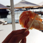 A close-up of a hand holding a big prawn between two chopsticks. Behind, blurred, you can see half of a seagull.