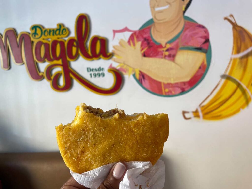 a close-up of a hand holding an arepa de huevo. Half of it is eaten. The arepa has a golden brown colour and is filled with ground meat