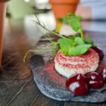 A round goat cheese cream on a black slap, topped with a red dust and green micro herbs. Next to it there are three dark red dollops of blackberry gel