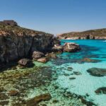 A view of a bay in Comino, with crustal clear water with a light shade of blue, through which you can see the rocks at the bottom of the sea. The cliffs are roughed, without much vegetation on them. A boat is passing in the distance