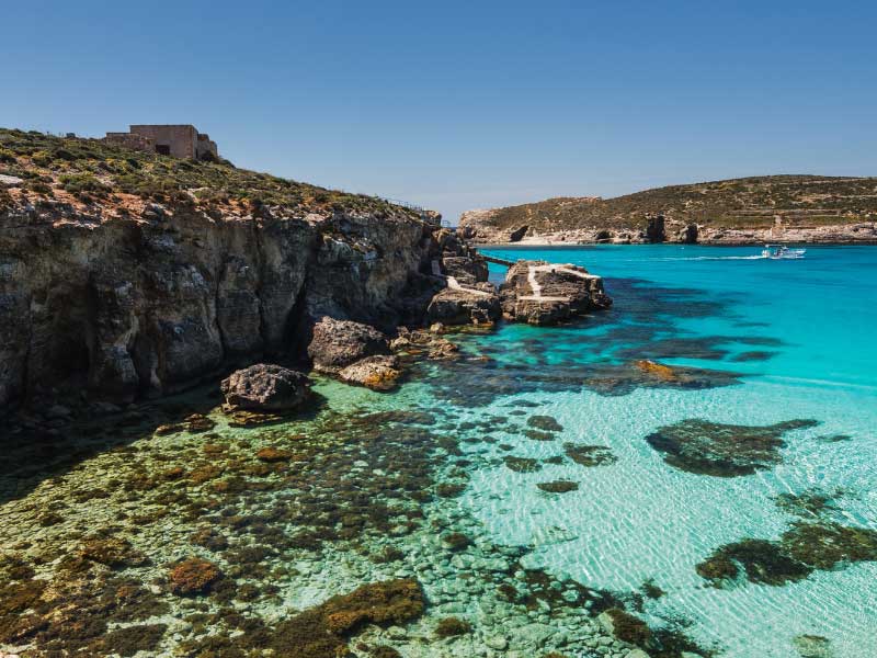 A view of a bay in Comino, with crustal clear water with a light shade of blue, through which you can see the rocks at the bottom of the sea. The cliffs are roughed, without much vegetation on them. A boat is passing in the distance