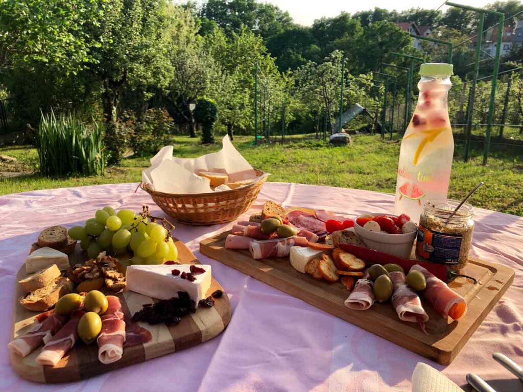 A charcuterie and cheese spread on two wooden boards, on a table outside, surrounded by nature.