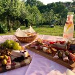 A charcuterie and cheese spread on two wooden boards, on a table outside, surrounded by nature.