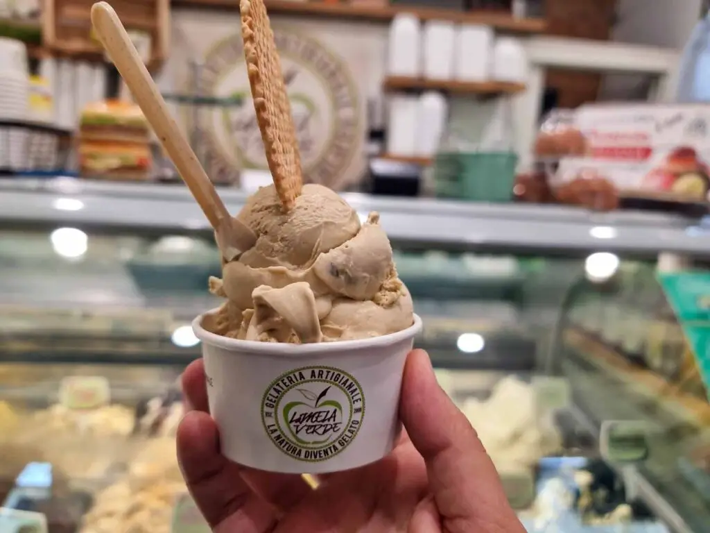 A white cup with the green logo of the gelateria on it. Inside there is a large quantity of gelato, which has a light brown colour and a round wafer on top