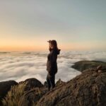 I am standing on a cliff, looking towards the scenery in front of me. There are fluffy clouds below me, filling the valley. The sky is orange, preparing for the sunrise. I am wearing blue jeans and a black jacket.