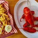 An oval plate on the right with an entire red lobster on it. On the left there is a basket with fries, a white paper bowl with coleslaw and a plastic cup filled with melted butter.