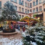 The market during the day time, with trees and the floor covered with snow. There are lit fairy lights everywhere.