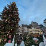 The Christmas tree in the Strasbourg Christmas market all lit up and decorated. It has large white baubles, circles with white snowflakes inside, yellow candle sticks and figurine of Santa Claus. It is night.