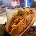 A close-up of my hand holding a kasekrainer sausage topped with mustard, crispy onions and slices of pickles