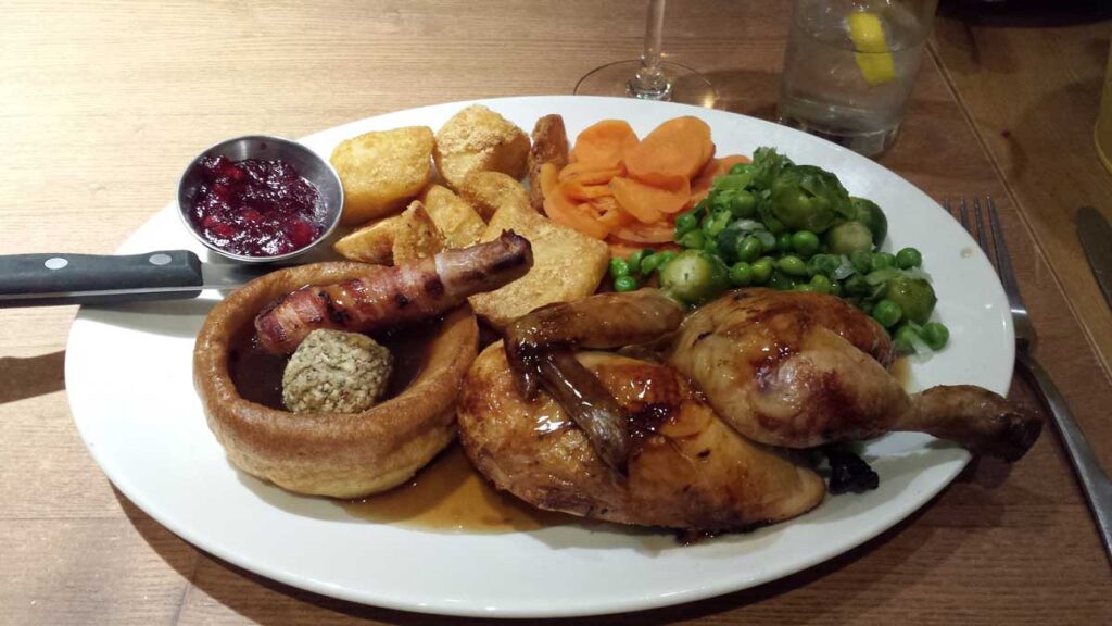 A plate of traditional roast dinner, with a roast piece of chicken, a Yorkshire pudding, a pig in blanket, stuffing, potatoes, greens, carrots, and a small bowl with cranberry sauce.