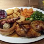 A plate of traditional roast dinner, with a roast piece of chicken, a Yorkshire pudding, a pig in blanket, stuffing, potatoes, greens, carrots, and a small bowl with cranberry sauce.
