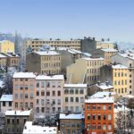 Pastel coloured 3 and 4 floors buildings with their flat rooftops covered in snow. The sky is blue above.