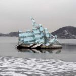 An artwork that resembles a ship, on the water. Towards the shore, there is ice on the water. The sky is grey.