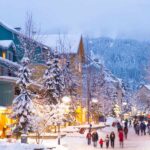 A street in Whistler covered with snow, with people strolling on it. The trees along the street have snow on them. In front there is a mountain with snow, in haze. There are buildings on the left hand side of the photo.