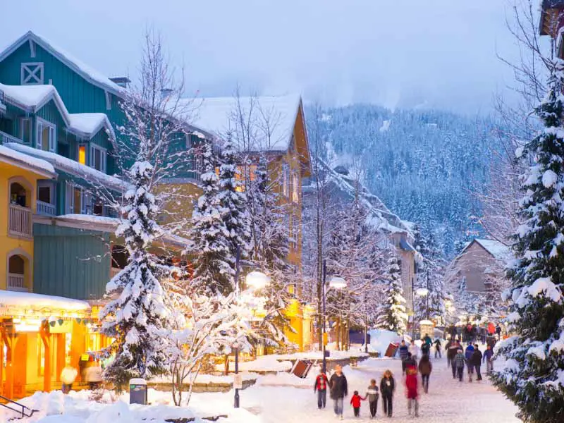 A street in Whistler covered with snow, with people strolling on it. The trees along the street have snow on them. In front there is a mountain with snow, in haze. There are buildings on the left hand side of the photo.