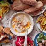 A table covered with a white lace tablecloth, with a lot of different dishes on it. There is a plate with bread, a bowl with chicken in a white sauce, fried chicken legs, a red tomato salad, a plate with fries, pickles, vegetable spreads.
