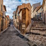 A narrow street in Port de Soller in Mallorca, between stone houses in a light brown colours. On the right there are stairs going up the hill.