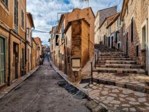 A narrow street in Port de Soller in Mallorca, between stone houses in a light brown colours. On the right there are stairs going up the hill.