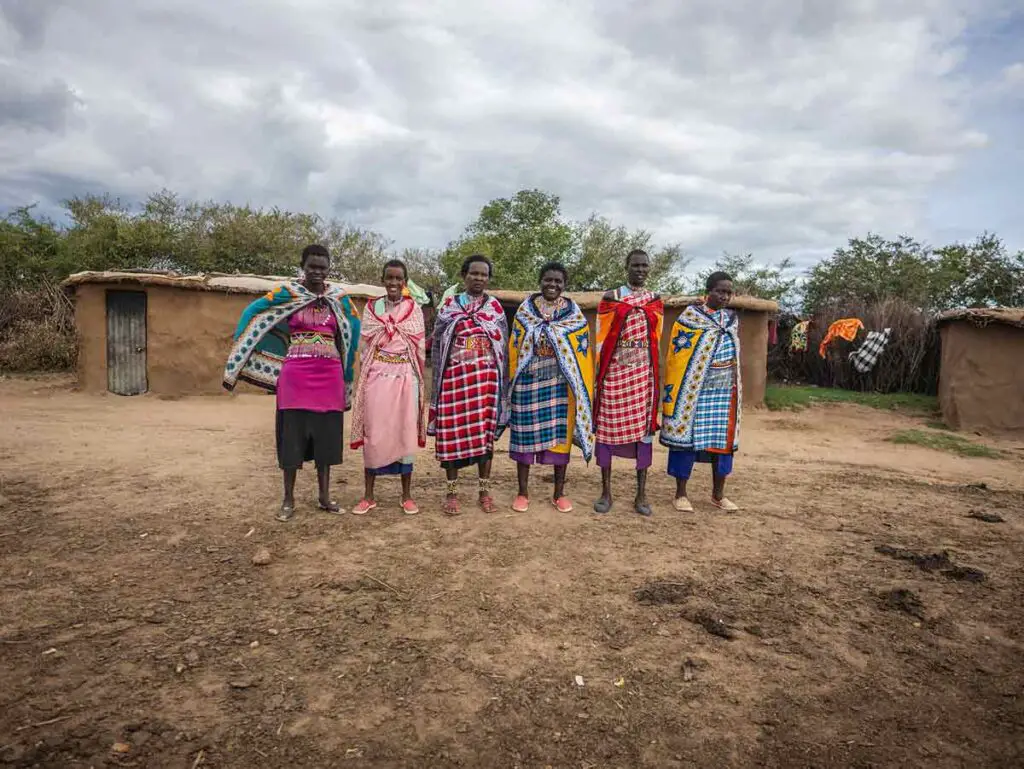 A group of Maasai women wearing very colourful traditional attire, standing in a line, smiling at the camera. Behind, you can see the typical Maasai mud houses. The sky above is very cloudy.