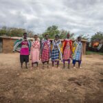 A group of Maasai women wearing very colourful traditional attire, standing in a line, smiling at the camera. Behind, you can see the typical Maasai mud houses. The sky above is very cloudy.