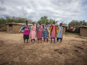A group of Maasai women wearing very colourful traditional attire, standing in a line, smiling at the camera. Behind, you can see the typical Maasai mud houses. The sky above is very cloudy.