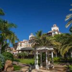 A beautiful serene corner in a garden, with a round pavilion in the middle, surrounded by palm trees and blue flower bushes. In the background you can see the hotel's building