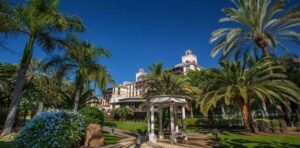 A beautiful serene corner in a garden, with a round pavilion in the middle, surrounded by palm trees and blue flower bushes. In the background you can see the hotel's building