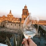 A glass of wine raised in front of the cathedral in mexico city, at dusk. The light is warm, as the sun just set.