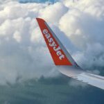 A closeup of the wing of an Easyjet plane, which is white and has the name of the company written on the tip of it, white on an orange background.