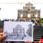A hand holding an old photo with a church facade on it, in front of the real church facade, in Macao. The postcard is black and white.