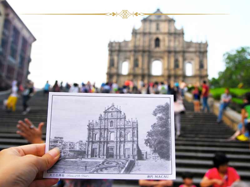 A hand holding an old photo with a church facade on it, in front of the real church facade, in Macao. The postcard is black and white.