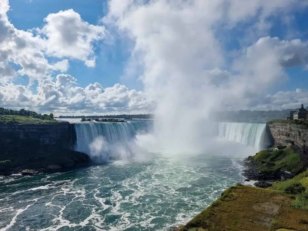 A photo of Niagara Falls on a sunny day, with just a few fluffy clouds on the sky. You can see the mist raising from the middle of the falls.