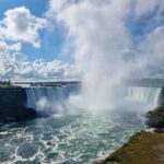 A photo of Niagara Falls on a sunny day, with just a few fluffy clouds on the sky. You can see the mist raising from the middle of the falls.
